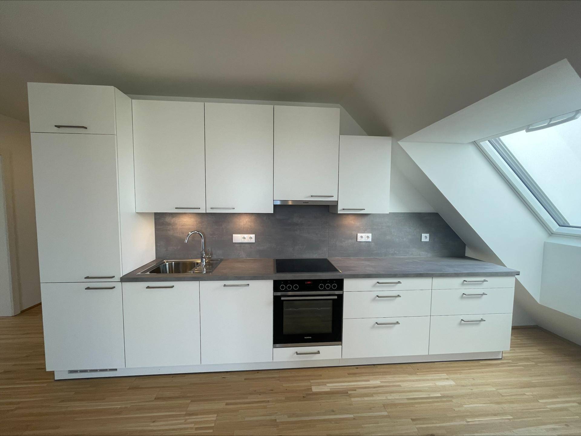 Moderne Einbauküche mit weißen Schränken, grauer Arbeitsplatte und integrierten Geräten unter Dachschrägen.