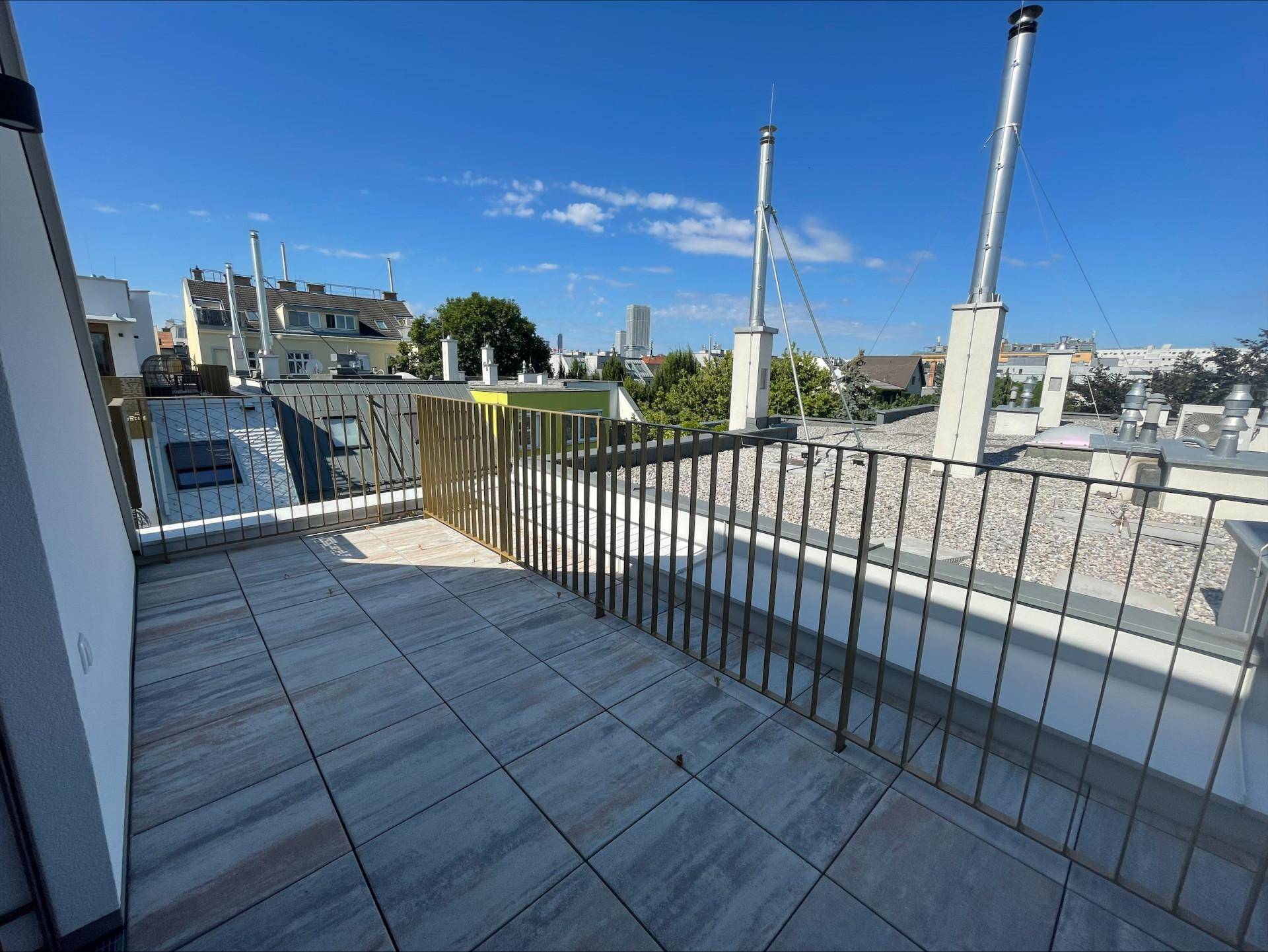 Sonniger Terrassenbereich mit Fliesenboden und schlichtem Metallgeländer, bietet weite Ausblicke über die Dächer.