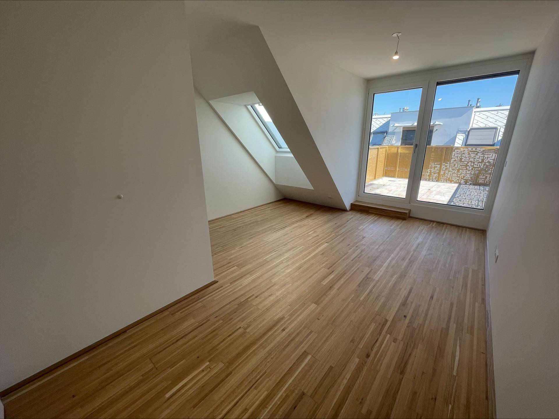 Geräumiges Zimmer mit Holzboden, Dachschräge und großer Glastür, die zu einem Außenbereich führt.