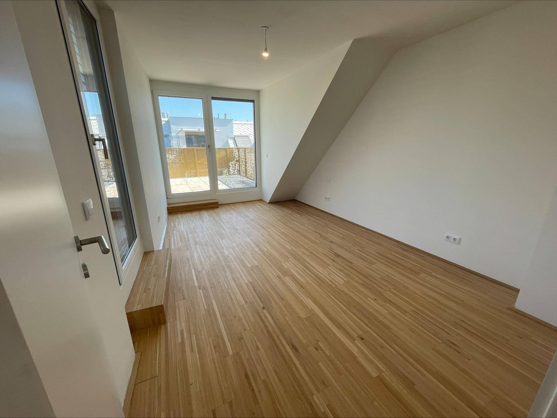 Helles Zimmer mit Holzboden, Dachschräge und Zugang zu einer Terrasse durch eine große Glastür.