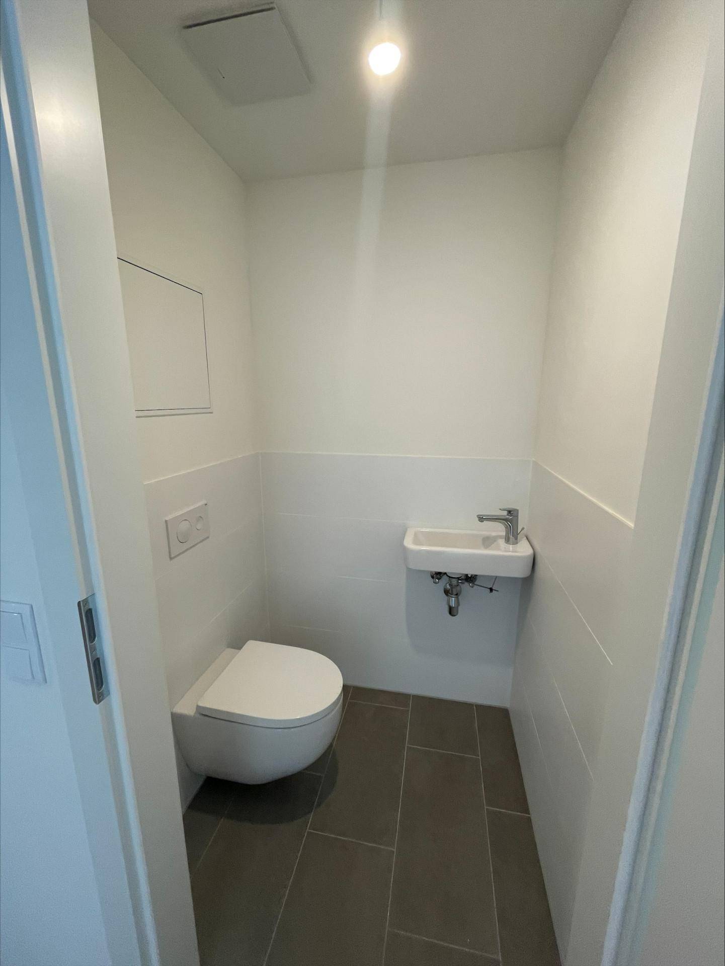 Modernes Gäste-WC mit wandhängender Toilette, kleinem Waschbecken und dunklen Bodenfliesen.