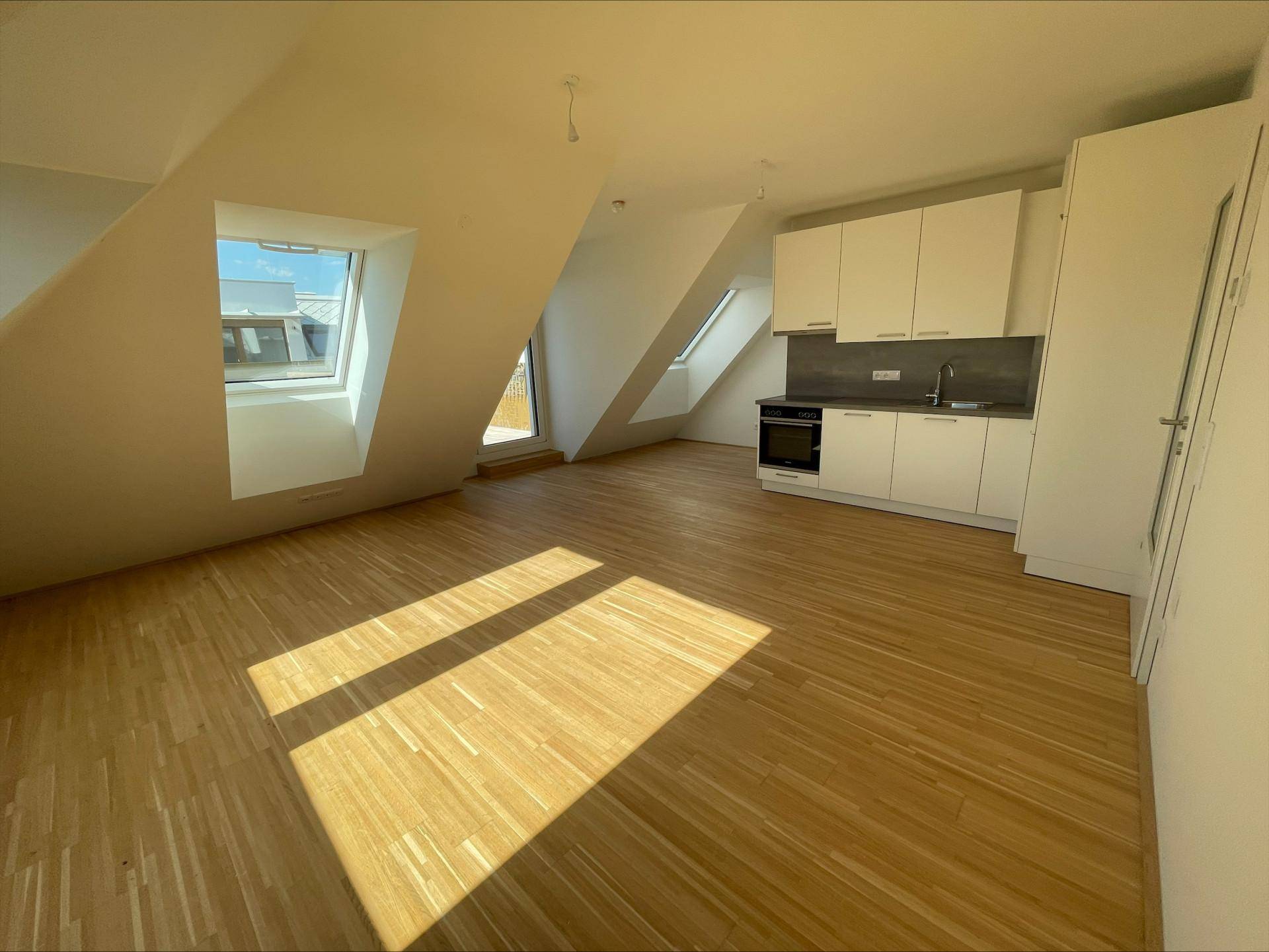 Heller Wohnraum mit Holzboden, Dachschrägen und integrierter Küchenzeile, durchflutet von natürlichem Licht.