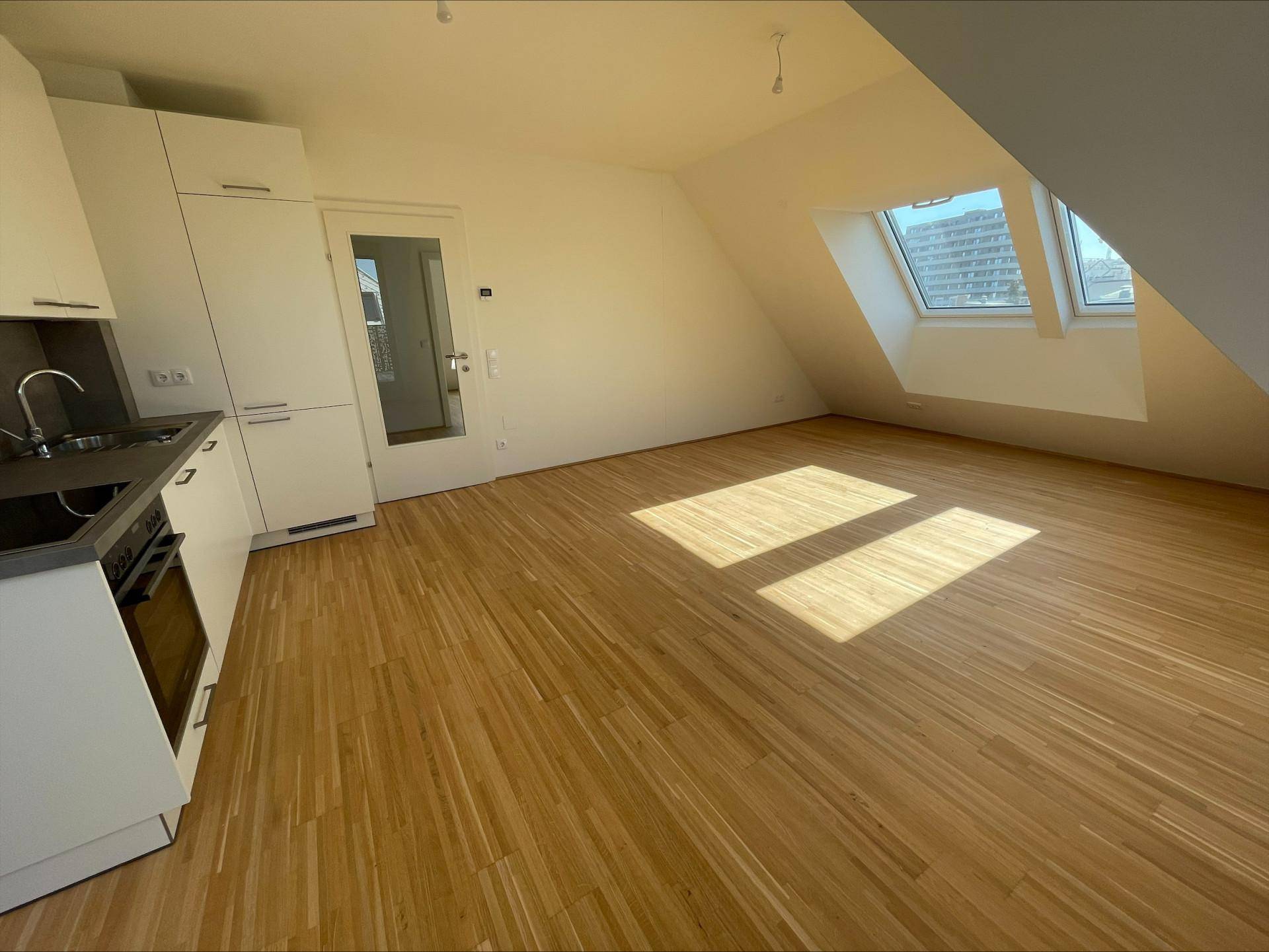 Lichtdurchfluteter Wohnbereich mit moderner Küchenzeile, Holzboden und Zugang zu einem weiteren Raum.