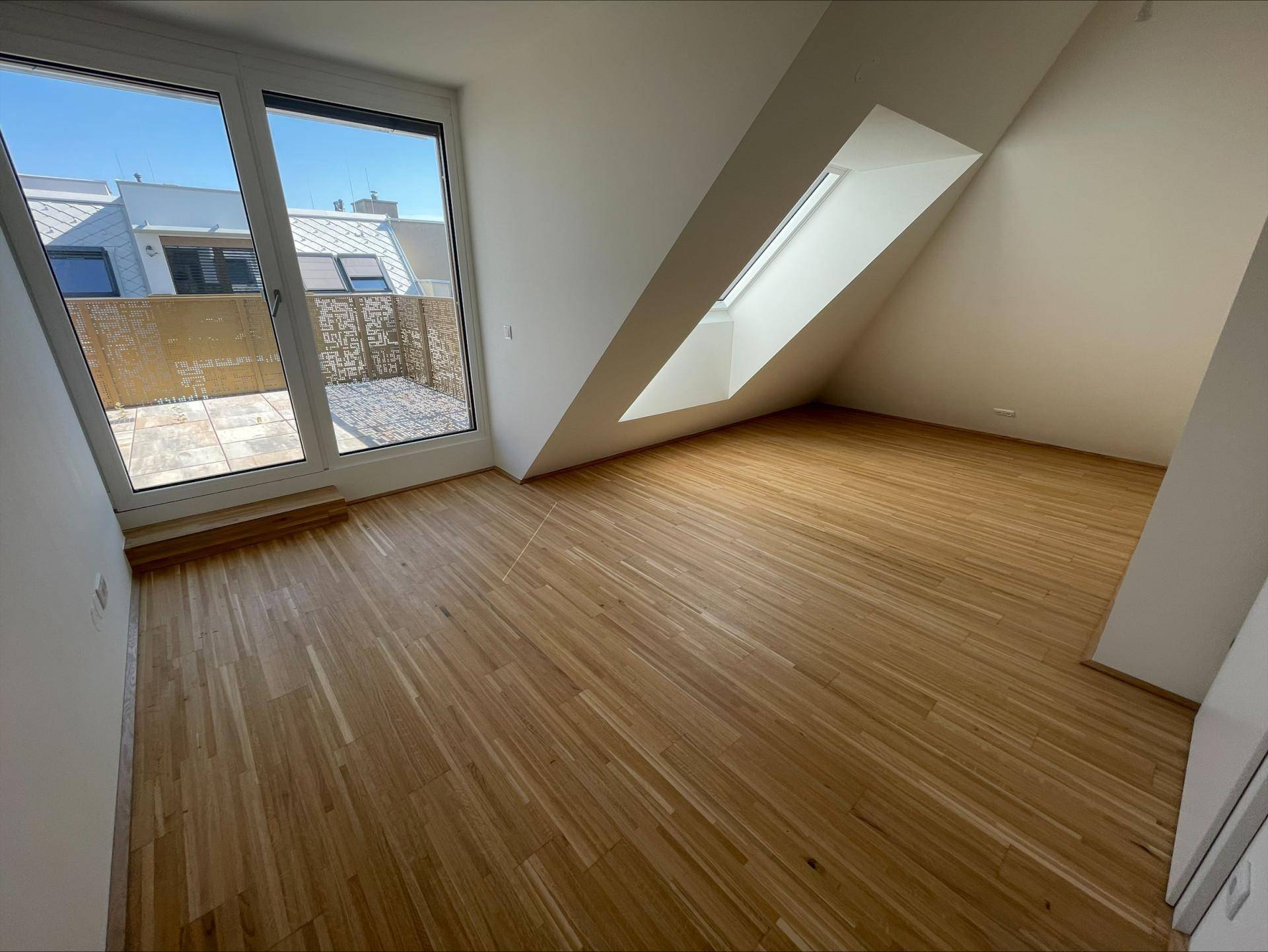 Lichtdurchflutetes Zimmer mit Holzboden, großer Terrassentür und Dachfenster, ideal für ein Schlafzimmer.