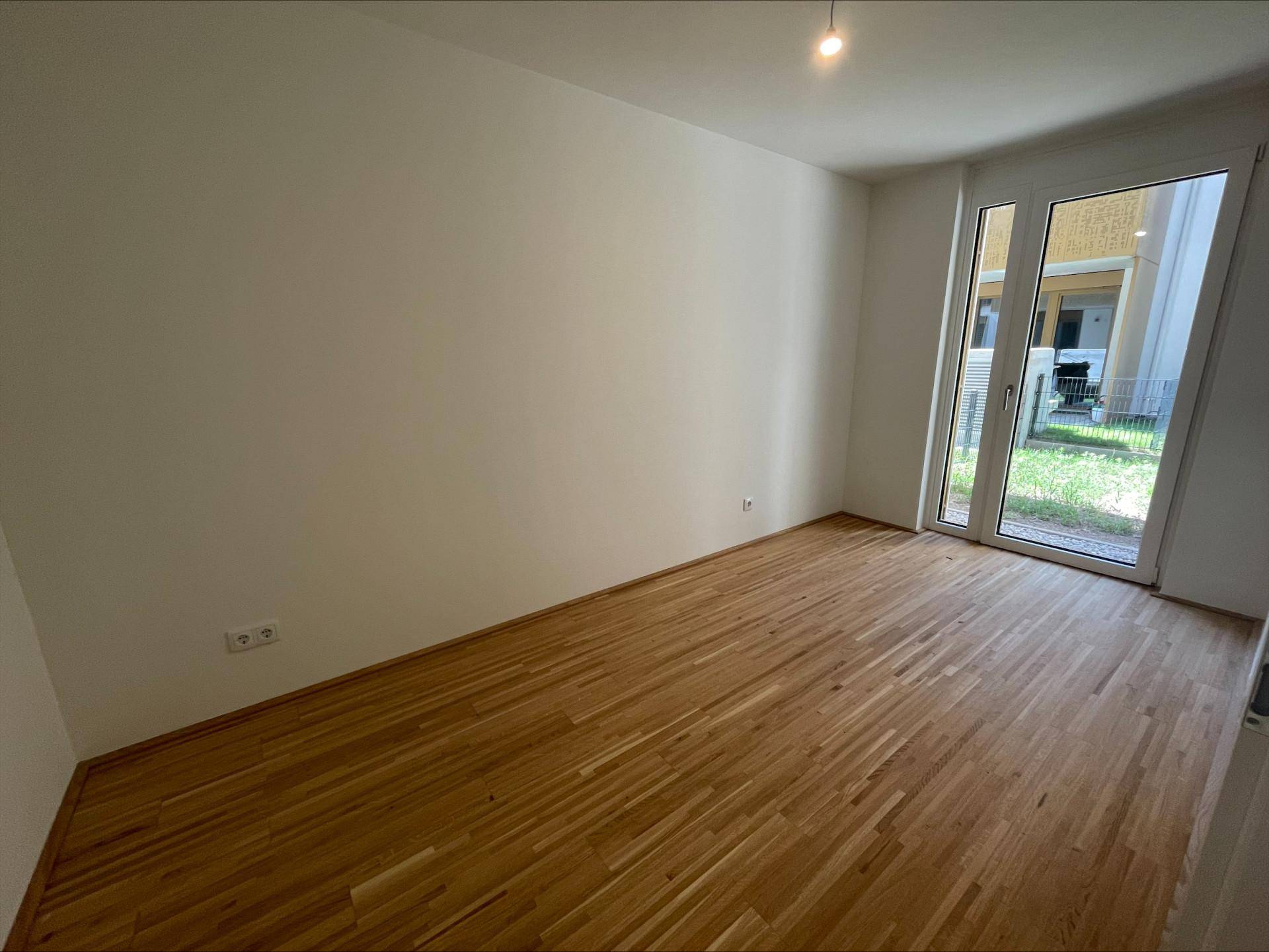 Leeres Zimmer mit Holzboden und großen Fenstern, die viel Tageslicht hereinlassen und Zugang zum Garten bieten.