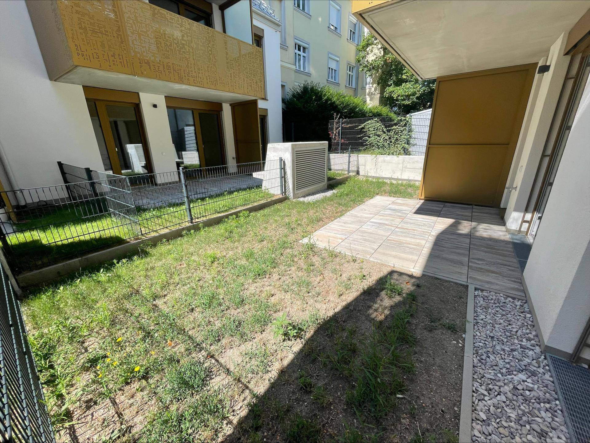 Kleiner Gartenbereich mit Rasenfläche und gepflasterter Terrasse, umgeben von einem Zaun.