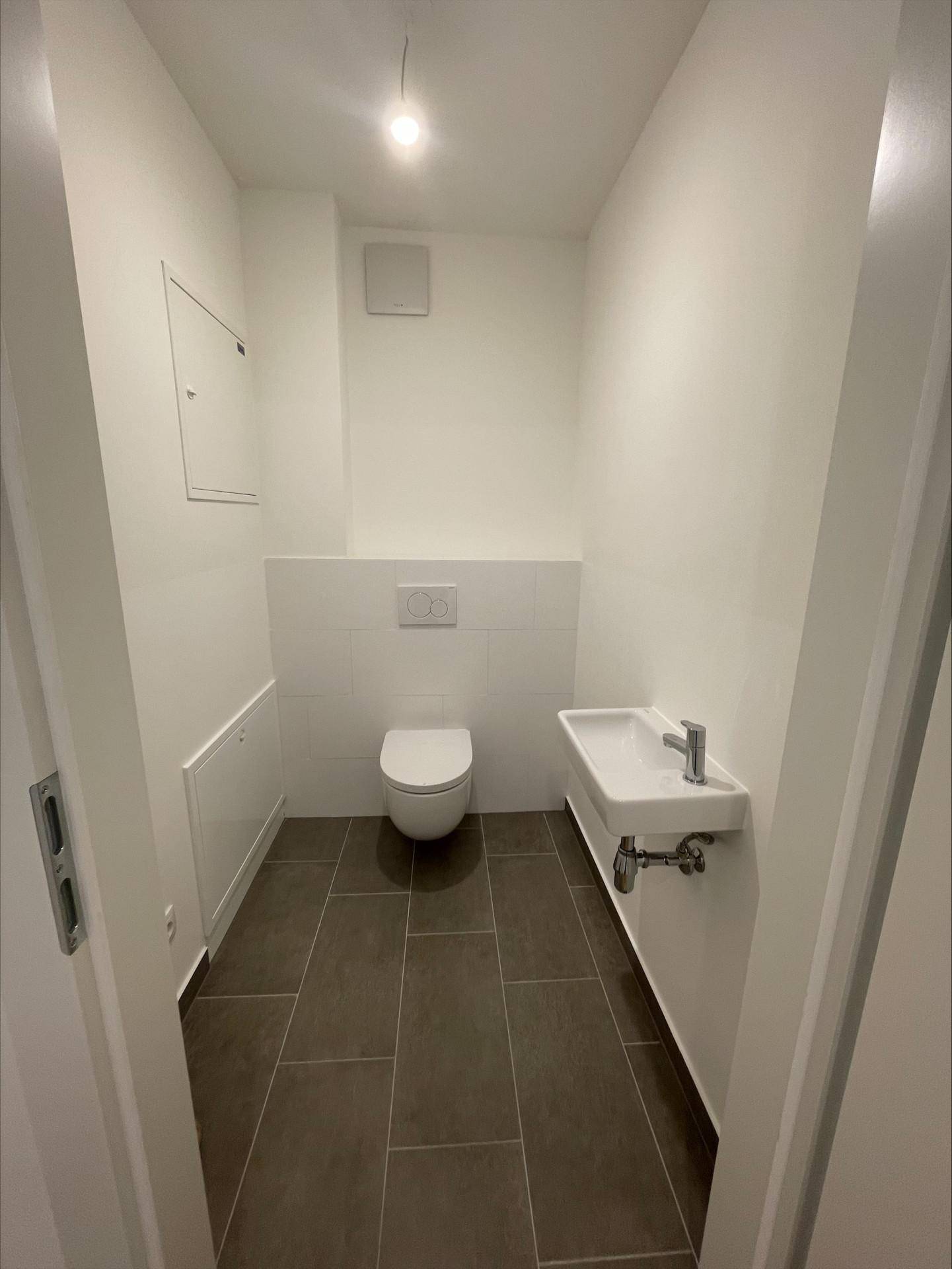 Separates Gäste-WC mit wandhängender Toilette, kleinem Waschbecken und dunklen Bodenfliesen.