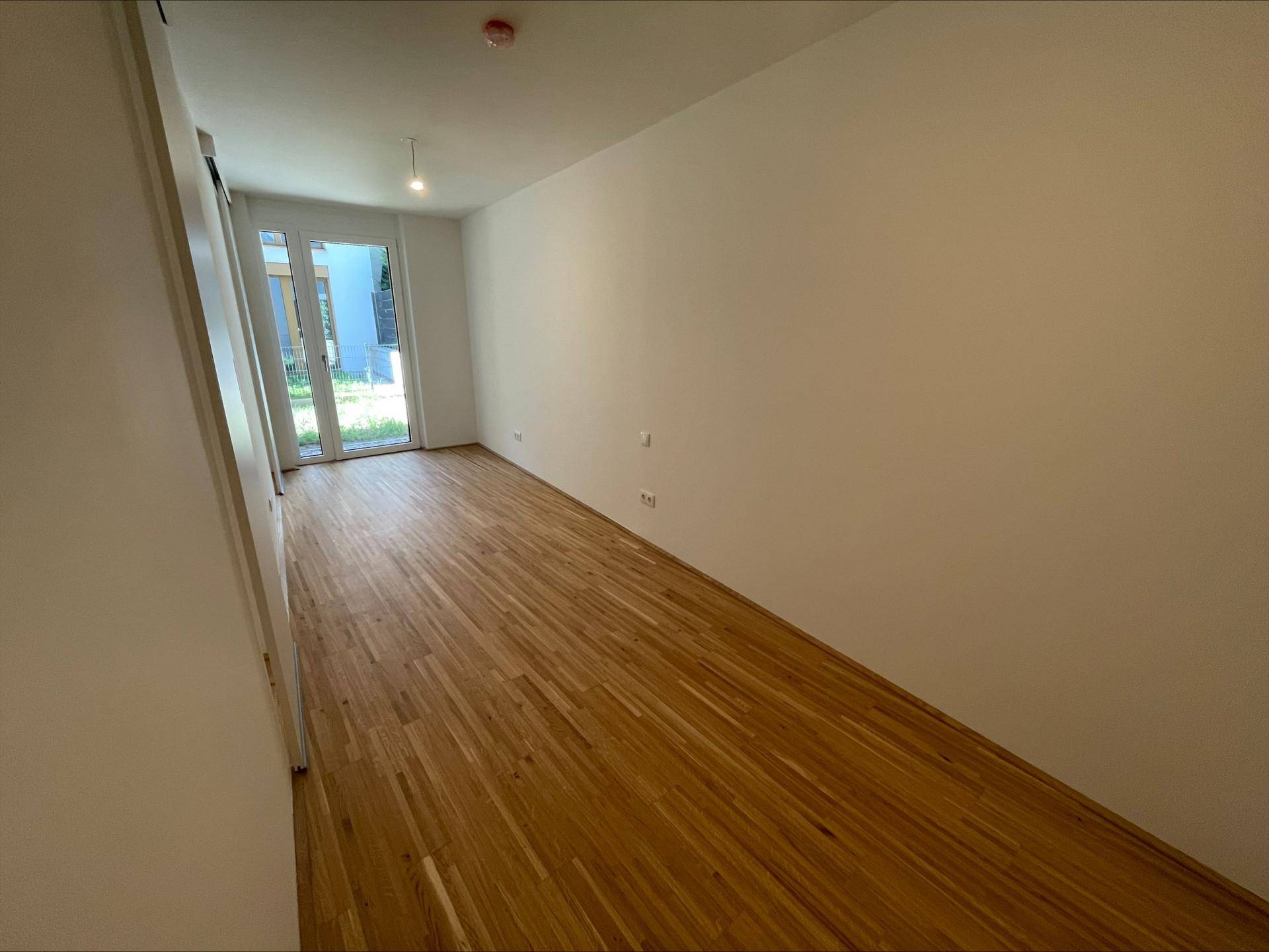 Geräumiges, leeres Zimmer mit Holzboden und Zugang zu einem Außenbereich durch große Fenster.