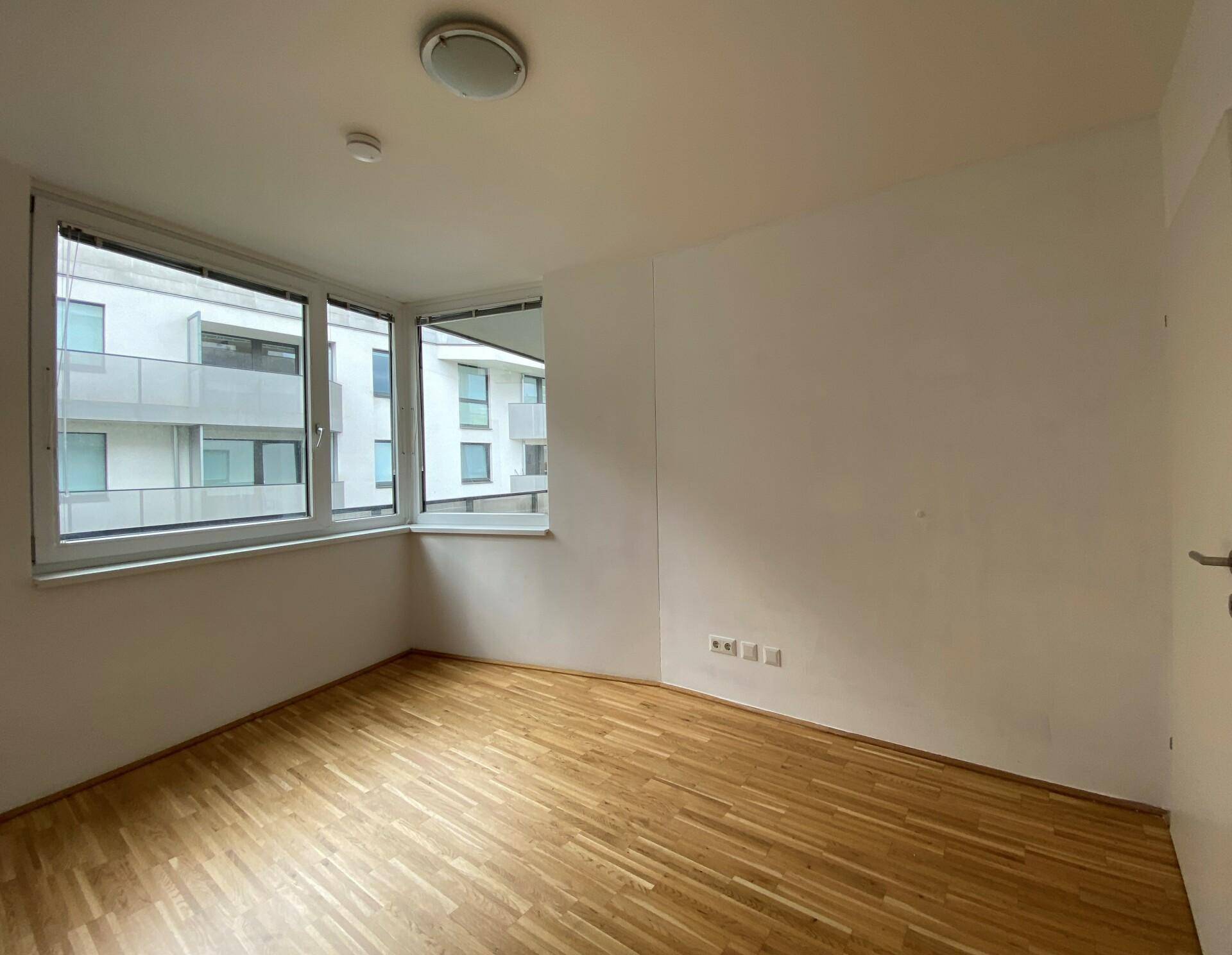 Helles Schlafzimmer mit Parkettboden und großem Fenster mit Blick auf die Nachbargebäude.