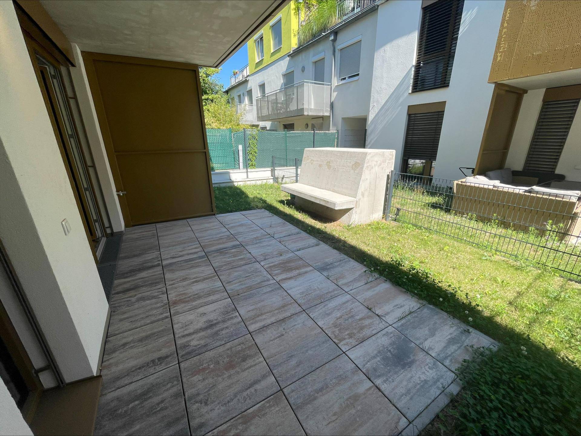 Sonniger Außenbereich mit gepflasterter Terrasse, Rasenfläche und Sitzbank.