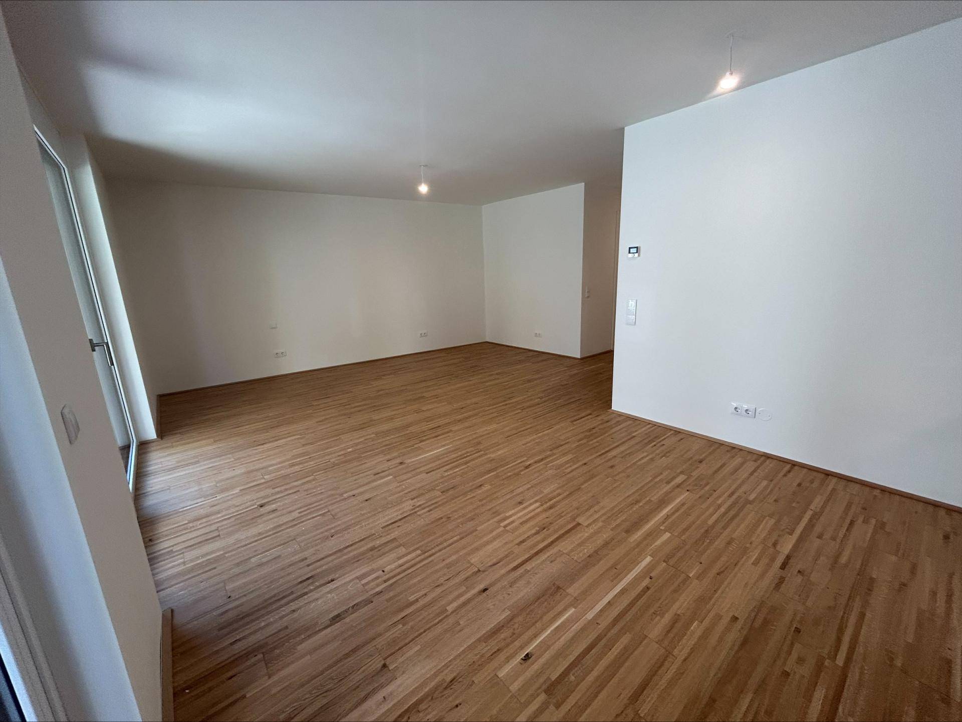 Geräumiges, leeres Wohnzimmer mit hellem Parkettboden und Zugang zu einem Außenbereich.