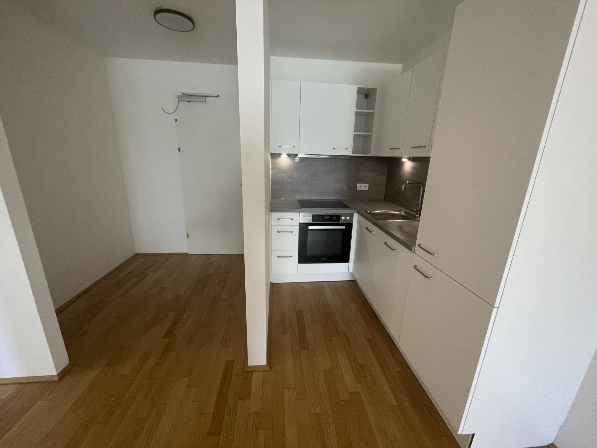Kleine, funktionale Küche mit weißen Schränken und Holzboden in einem leeren Raum.
