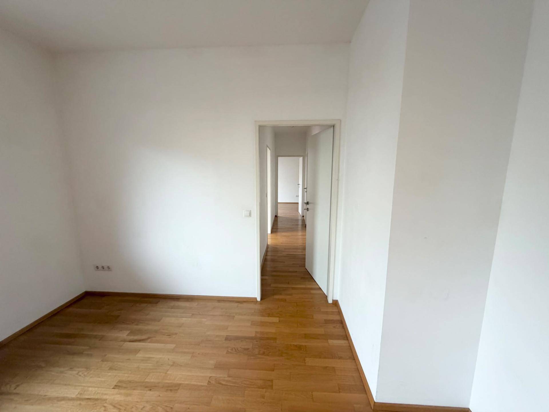 Leeres Schlafzimmer mit Parkettboden und Zugang zu einem Flur mit weiteren Zimmern, hell und freundlich.