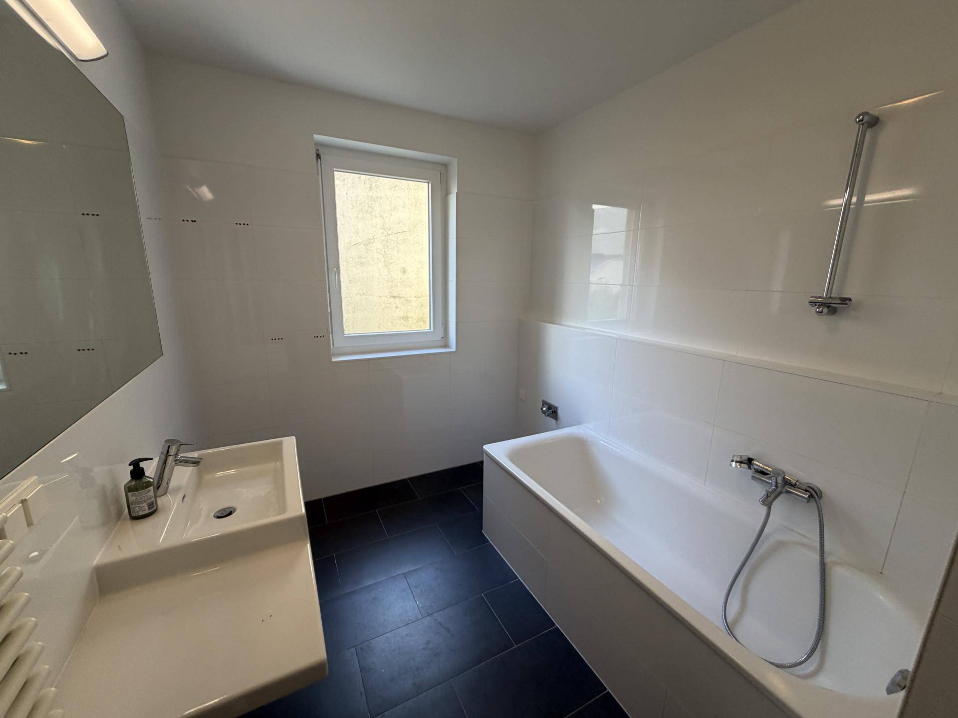 Helles Badezimmer mit Badewanne, Waschbecken, Fenster und dunklen Bodenfliesen.
