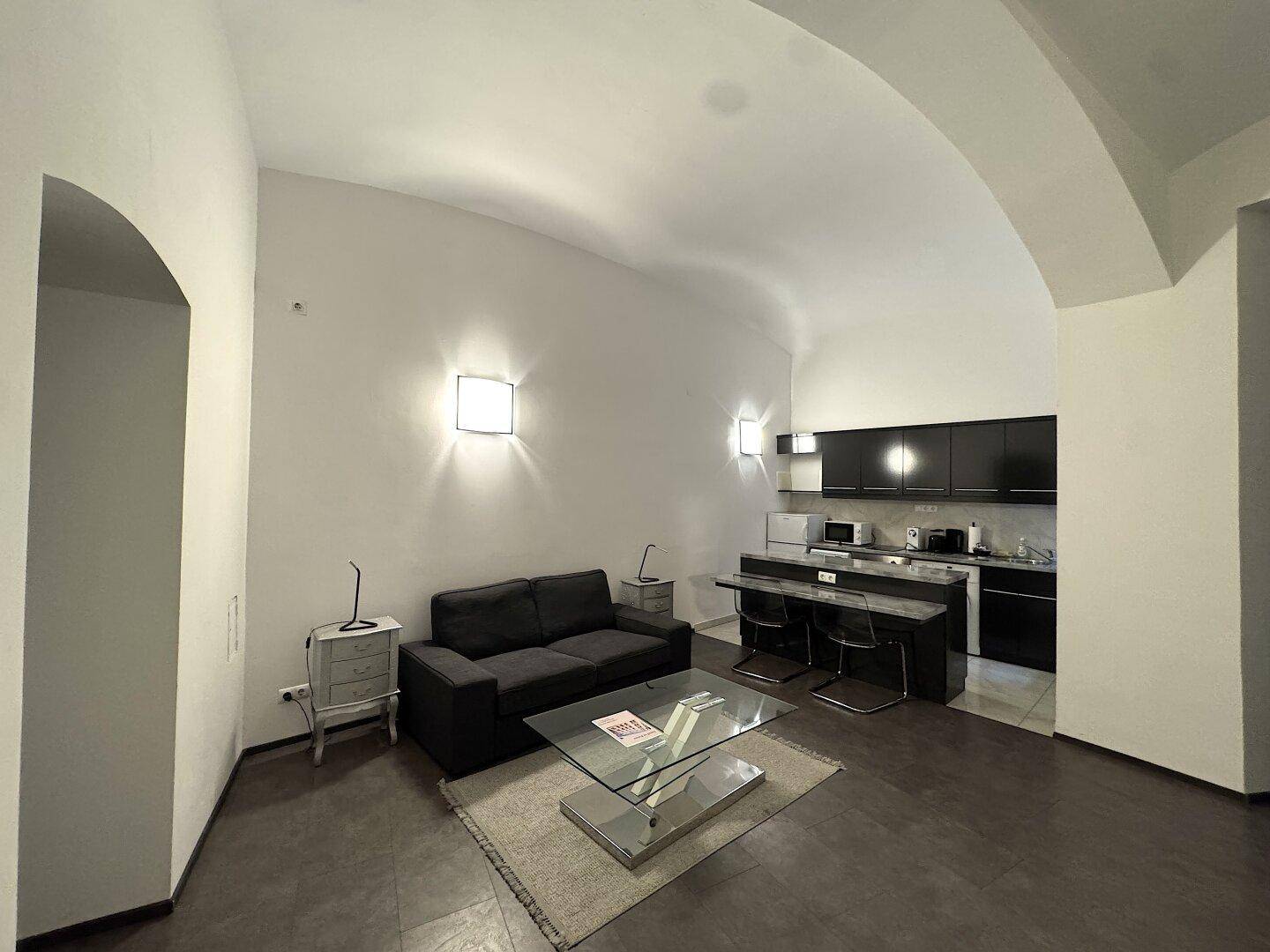 Geräumiger Wohnbereich mit bequemer Couch, Glastisch und integrierter Küchenzeile.