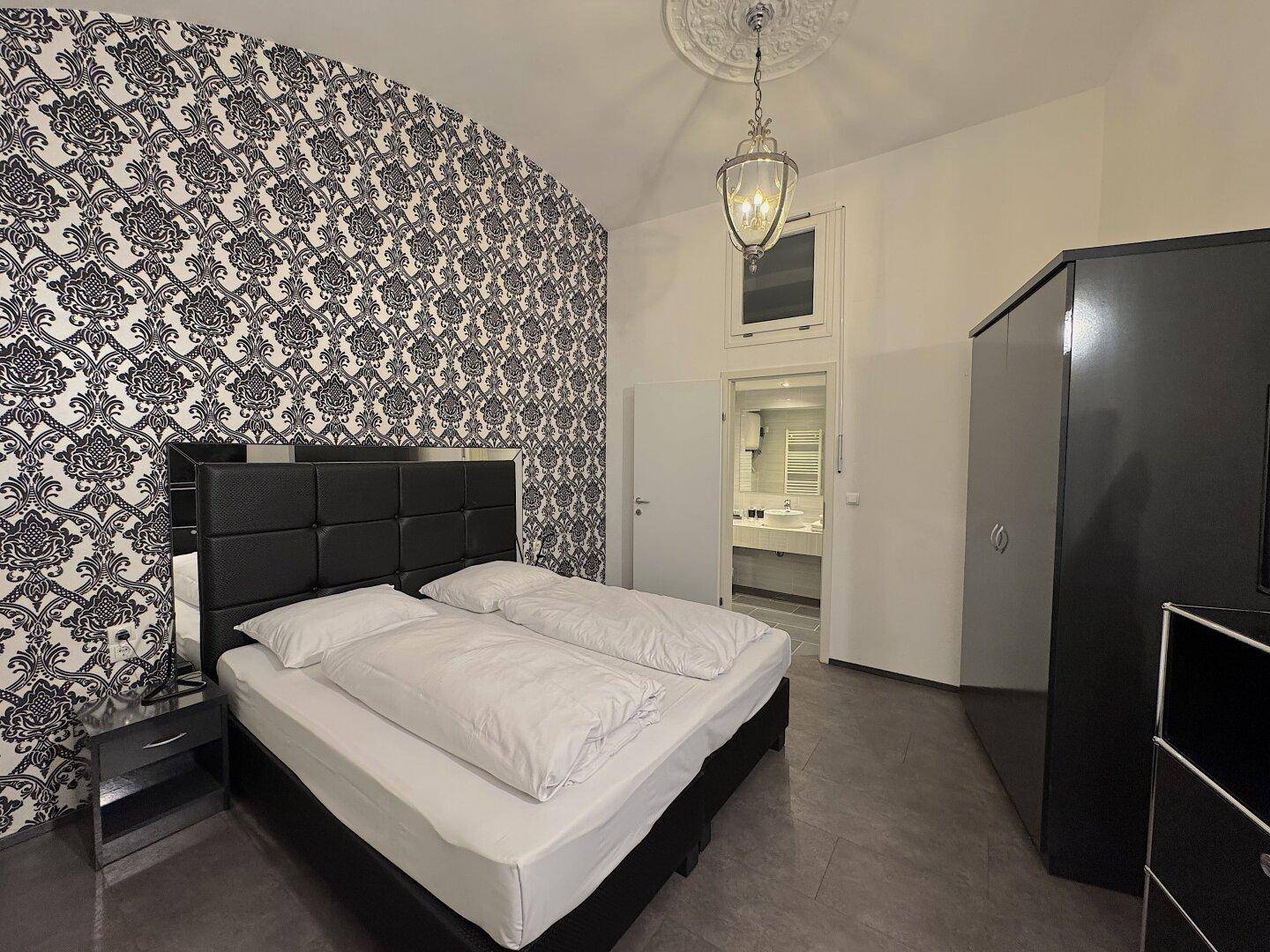 Schlafzimmer mit gemusterter Tapete, Doppelbett und direktem Zugang zum Badezimmer.