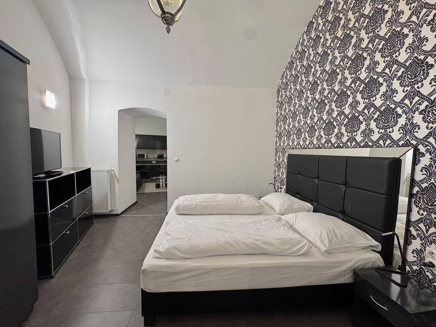 Schlafzimmer mit auffälliger gemusterter Tapete, Doppelbett und dunklen Möbeln.