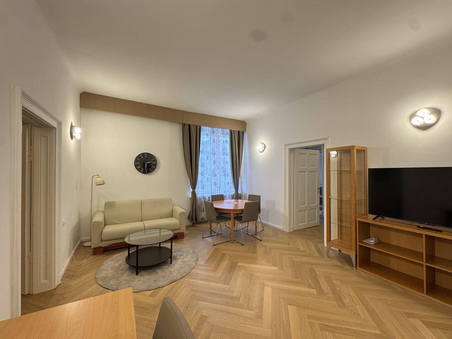 Wohn-Essbereich mit hellem Fischgrätparkett, gemütlichem Sofa und Fernseher.