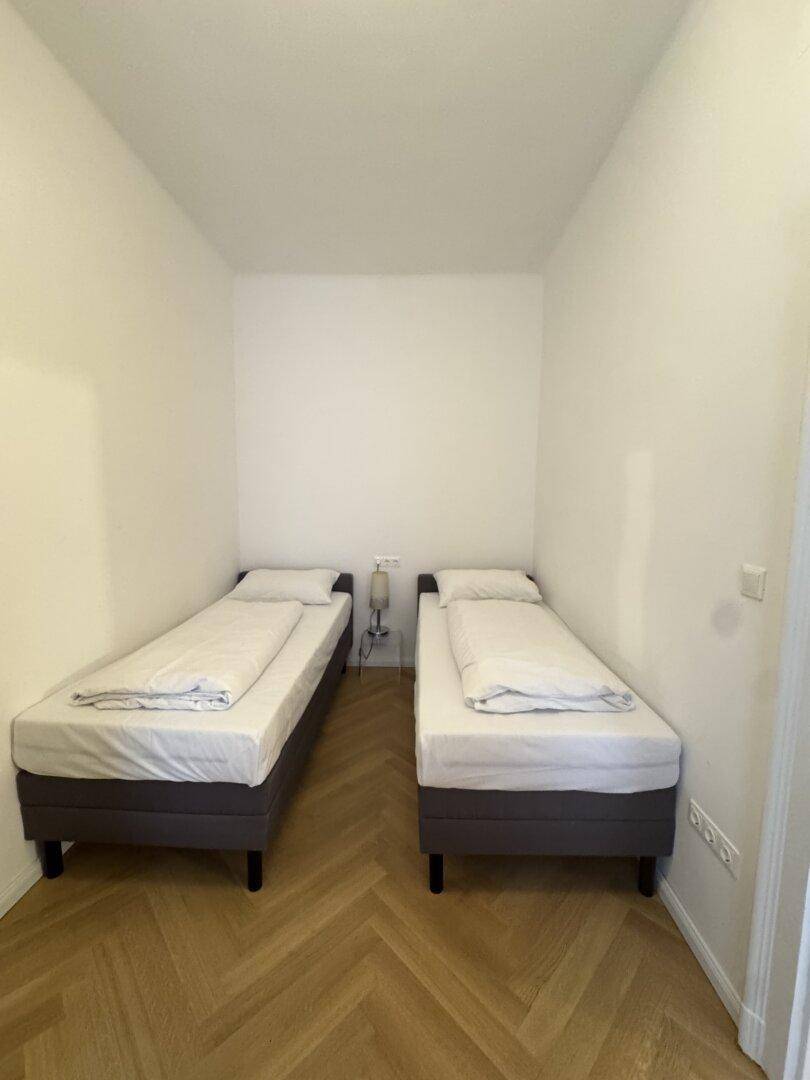Schlafzimmer mit zwei Einzelbetten, hellem Fischgrätparkett und weißen Wänden.