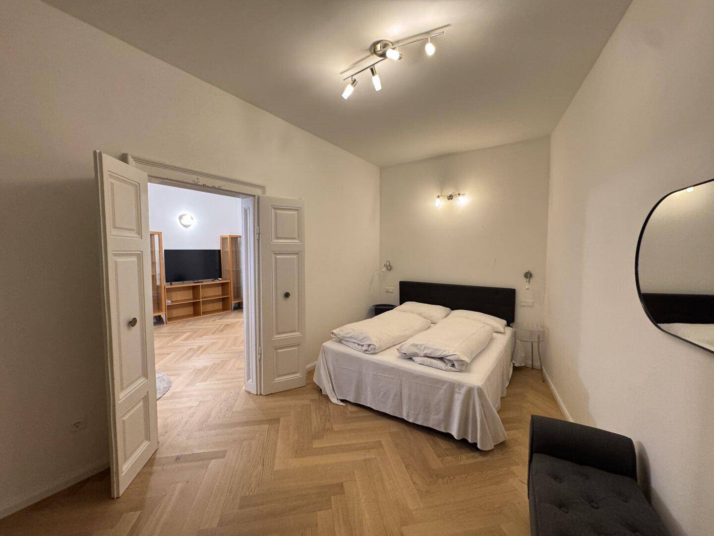 Schlafzimmer mit Doppelbett, ovalem Spiegel und Blick in den Wohnbereich.