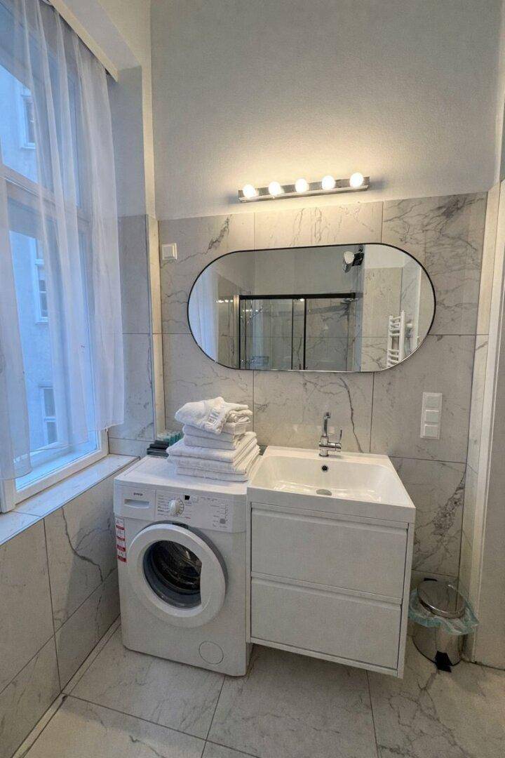Badezimmer mit Waschmaschine, Waschbecken, Spiegel und Fenster, gefliest in Marmoroptik.