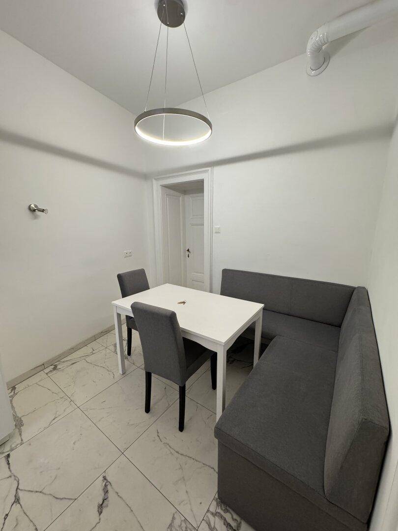 Essbereich in der Küche mit weißem Tisch, grauer Sitzbank und Fliesenboden.