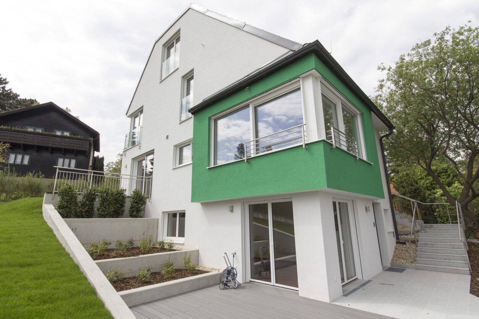 Moderne Hausfassade mit grünen Akzenten und großen Fenstern, umgeben von einer gepflegten Außenanlage.