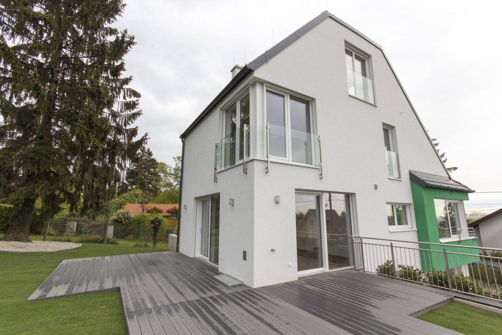 Moderne Hausfassade mit großer Holzterrasse und Zugang zum Garten, umgeben von viel Grün.