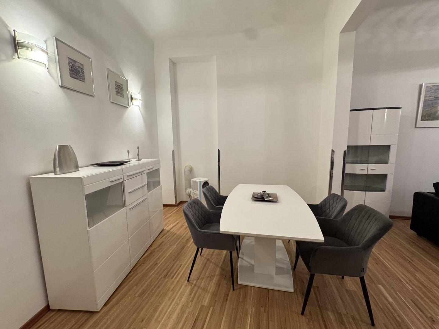Moderner Essbereich mit weißem Tisch, grauen Stühlen und einem Sideboard, angrenzend an den Wohnbereich.