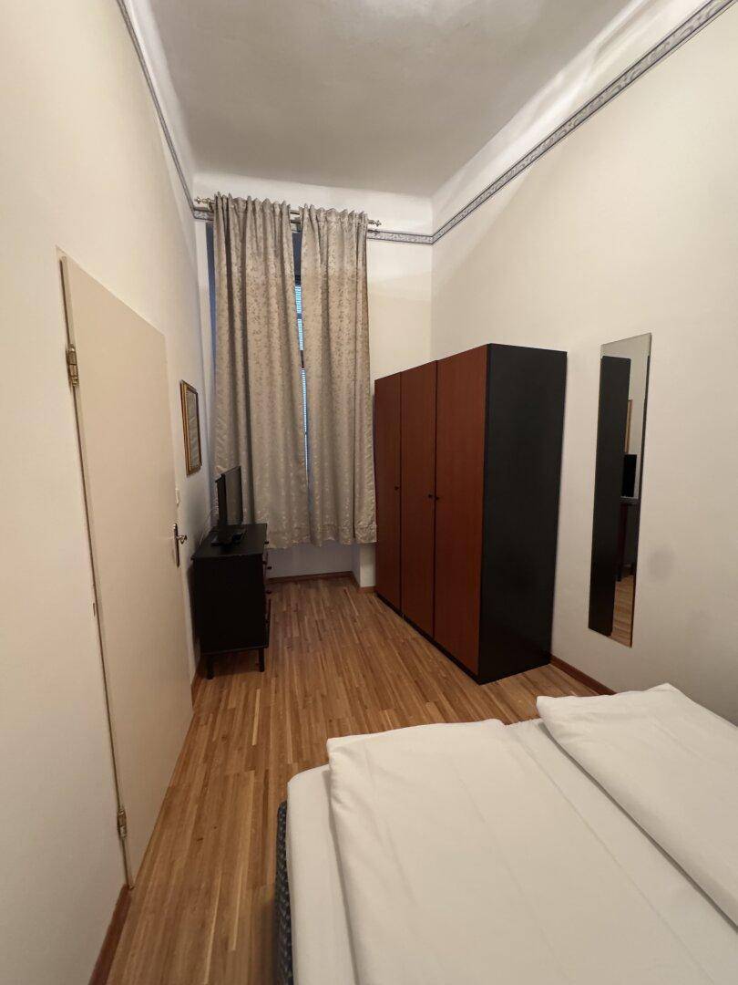 Schlafzimmer mit Holzparkett, Kleiderschrank und einem Fenster mit Vorhängen, das Tageslicht hereinlässt.