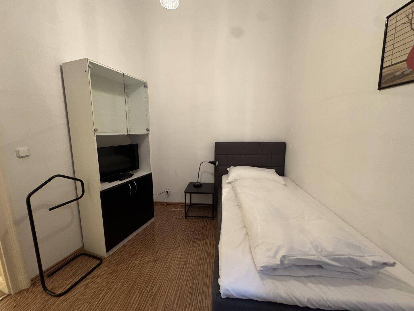 Schlafzimmer mit Einzelbett, kleinem Fernseher und einem Regal an der Wand.