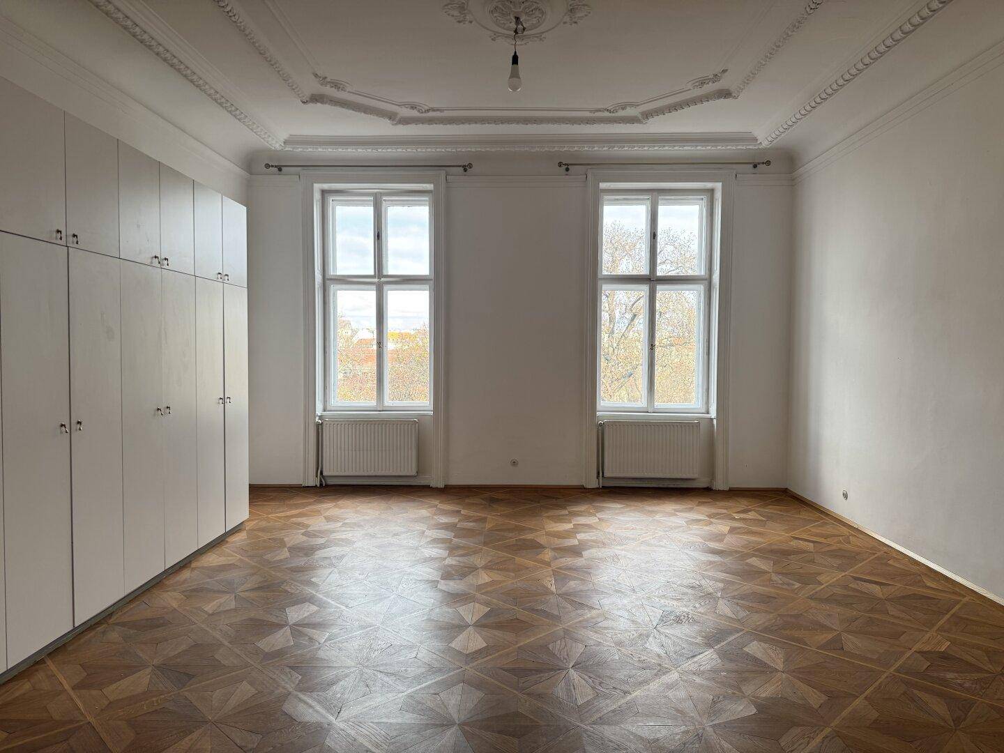 Heller Raum mit Fischgrätparkett, hohen Decken und großen Fenstern, die einen Blick ins Grüne bieten.