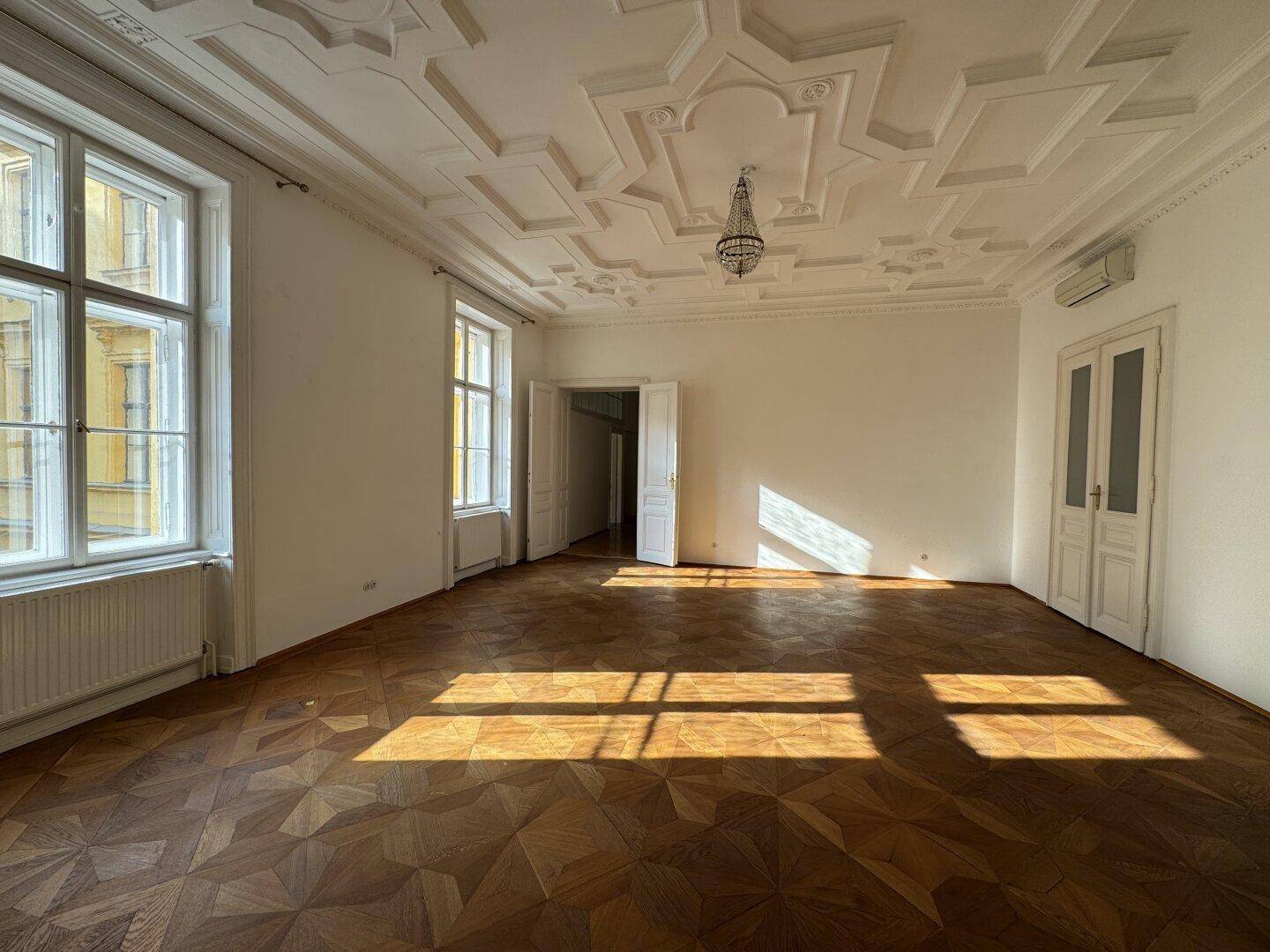 Großzügiges Wohnzimmer mit dekorativer Stuckdecke, Parkettboden und großen Fenstern, die viel Licht hereinlassen.