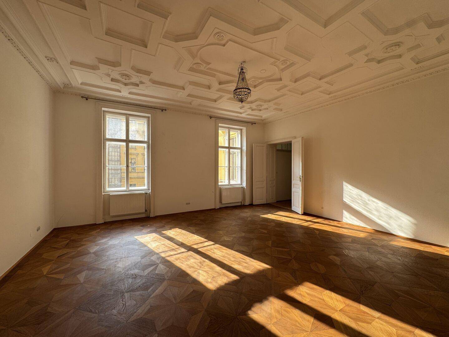 Geräumiges Wohnzimmer mit kunstvoller Stuckdecke, Fischgrätparkett und viel natürlichem Lichteinfall.
