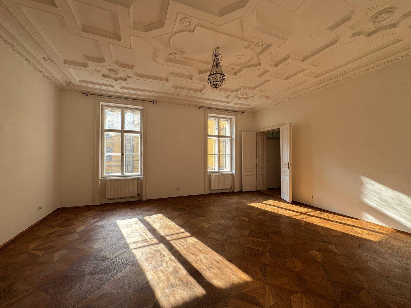Geräumiges Wohnzimmer mit kunstvoller Stuckdecke, Fischgrätparkett und viel natürlichem Lichteinfall.