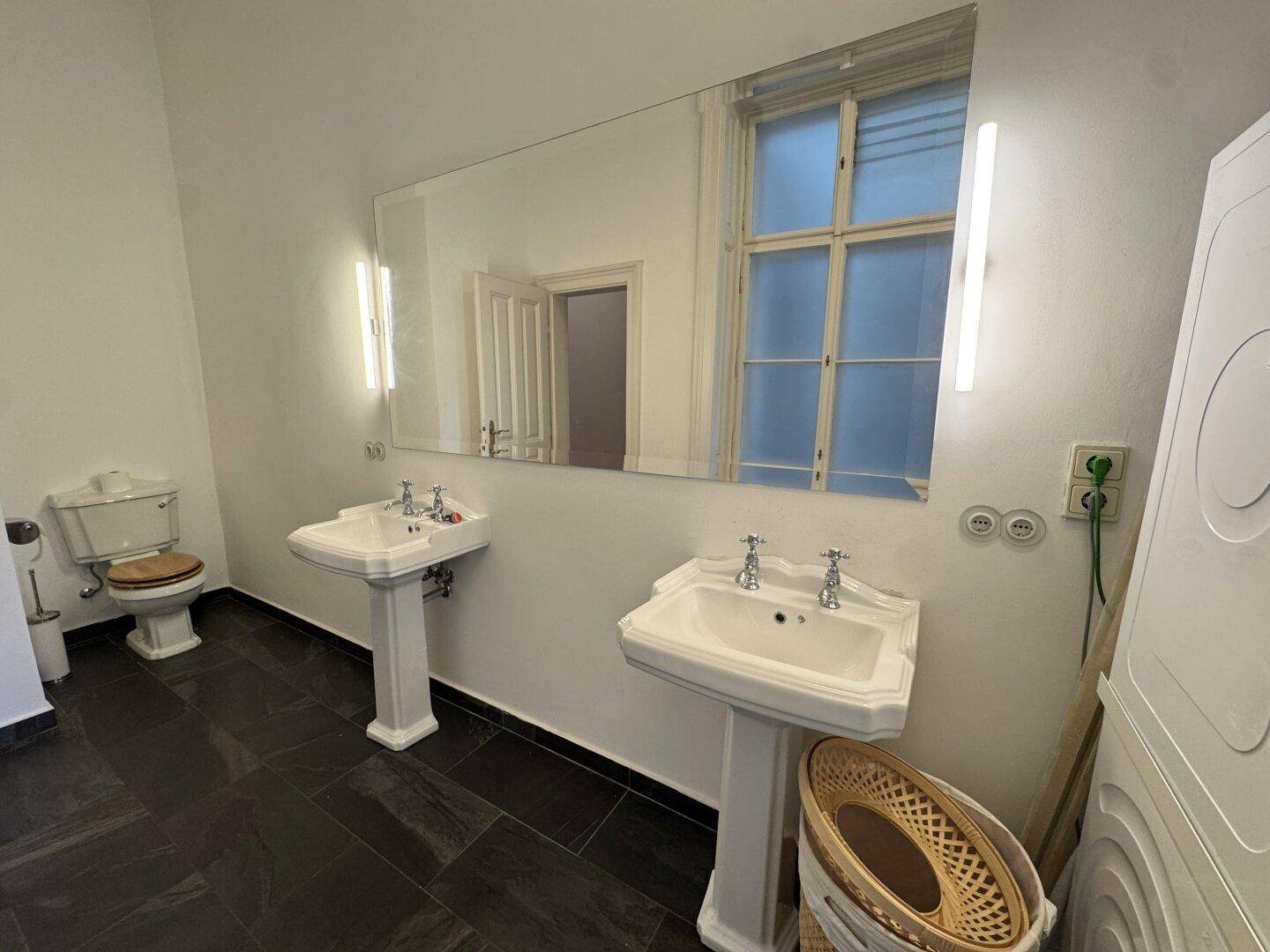 Badezimmer mit zwei Waschbecken, großem Spiegel und dunklen Fliesen, inklusive Waschmaschine.