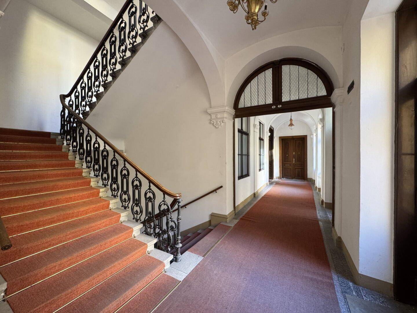 Elegantes Treppenhaus mit schmiedeeisernem Geländer, rotem Teppich und hohen Decken in einem Altbau.