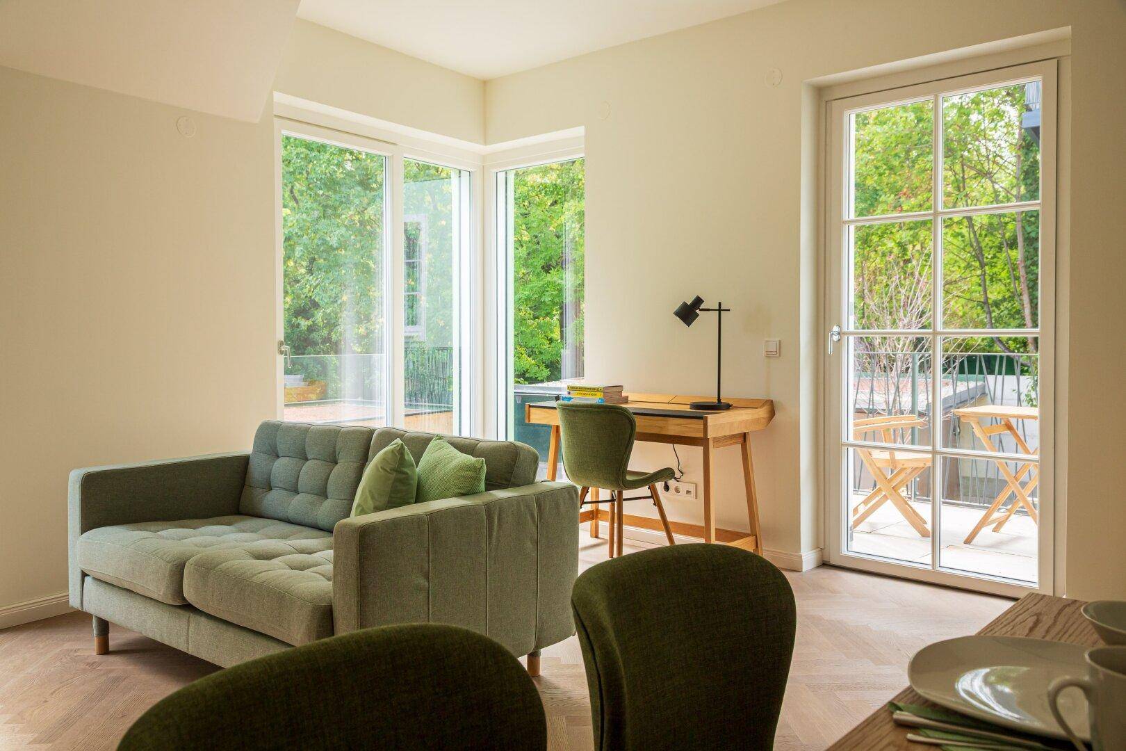 Gemütlicher Wohnbereich mit moderner Couch, Schreibtisch und großen Fenstern mit Blick ins Grüne.