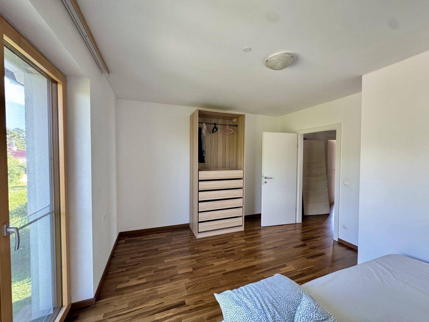 Helles Schlafzimmer mit Holzboden, Einbauschrank und großem Fenster mit Blick ins Grüne.