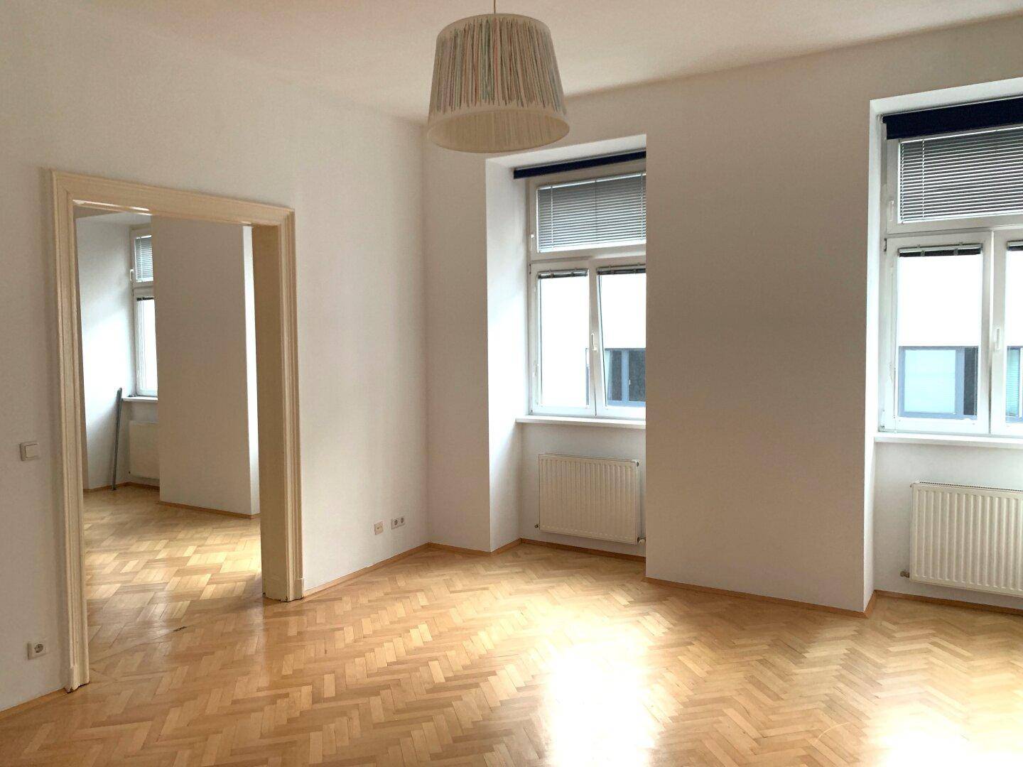Helles Zimmer mit Fischgrätparkettboden und zwei Fenstern, offen zu einem weiteren Raum.