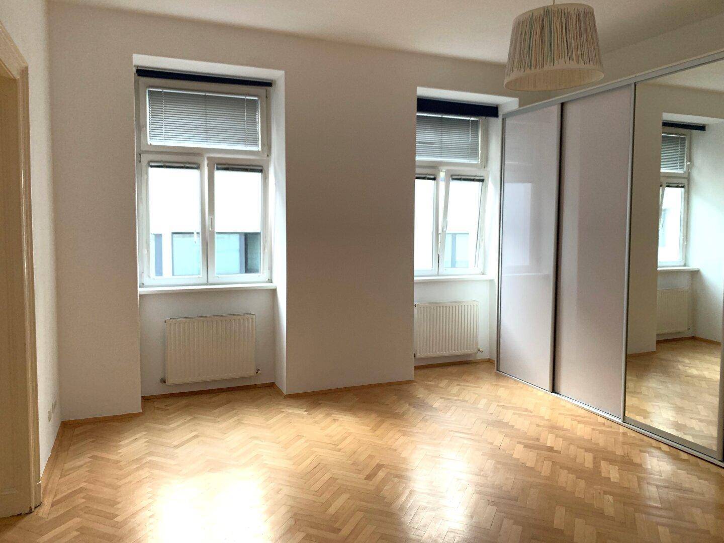 Geräumiges Schlafzimmer mit Fischgrätparkett, zwei Fenstern und einem großen Spiegelschrank.