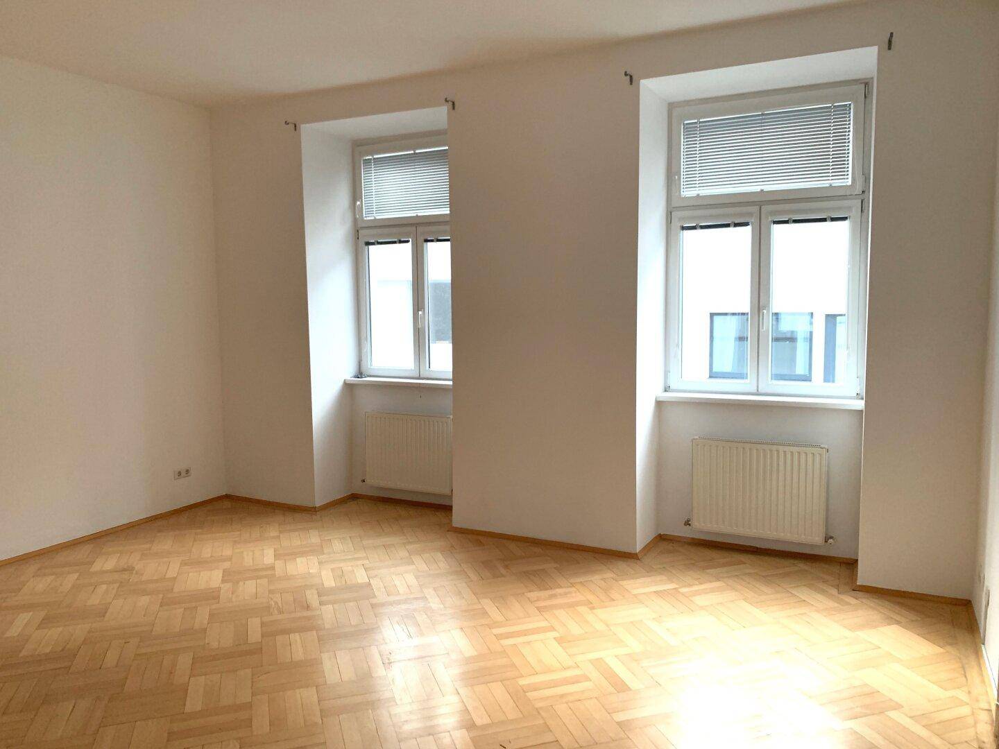 Geräumiges Zimmer mit Fischgrätparkettboden und zwei großen Fenstern, gut beleuchtet.