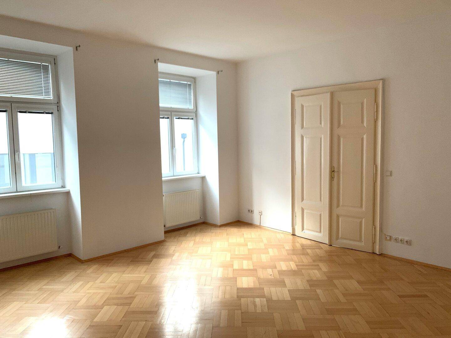 Helles Zimmer mit Fischgrätparkett, zwei Fenstern und einer geschlossenen Doppeltür.
