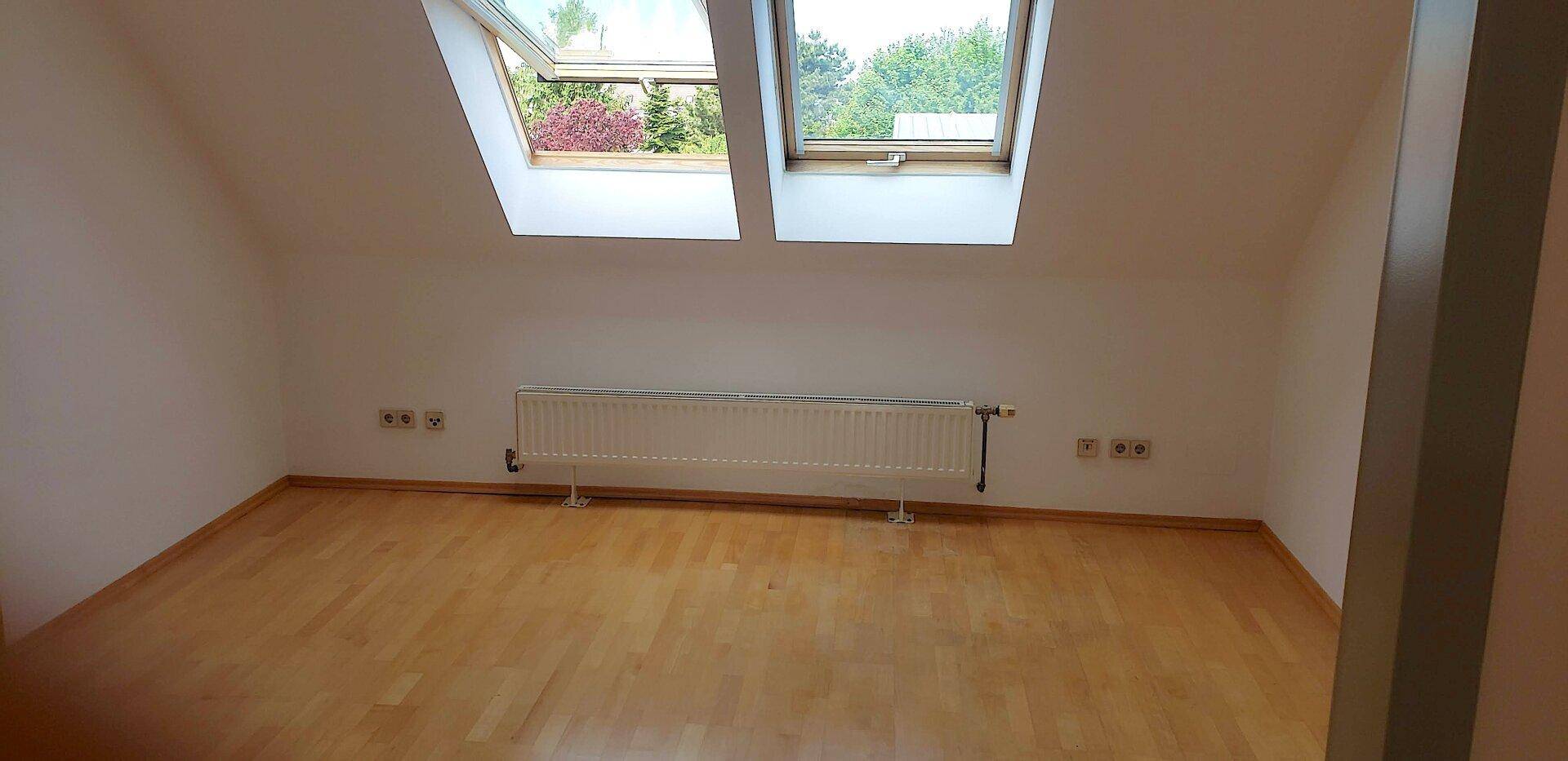 Helles Dachzimmer mit Holzfußboden, Dachschrägen und zwei großen Dachfenstern.