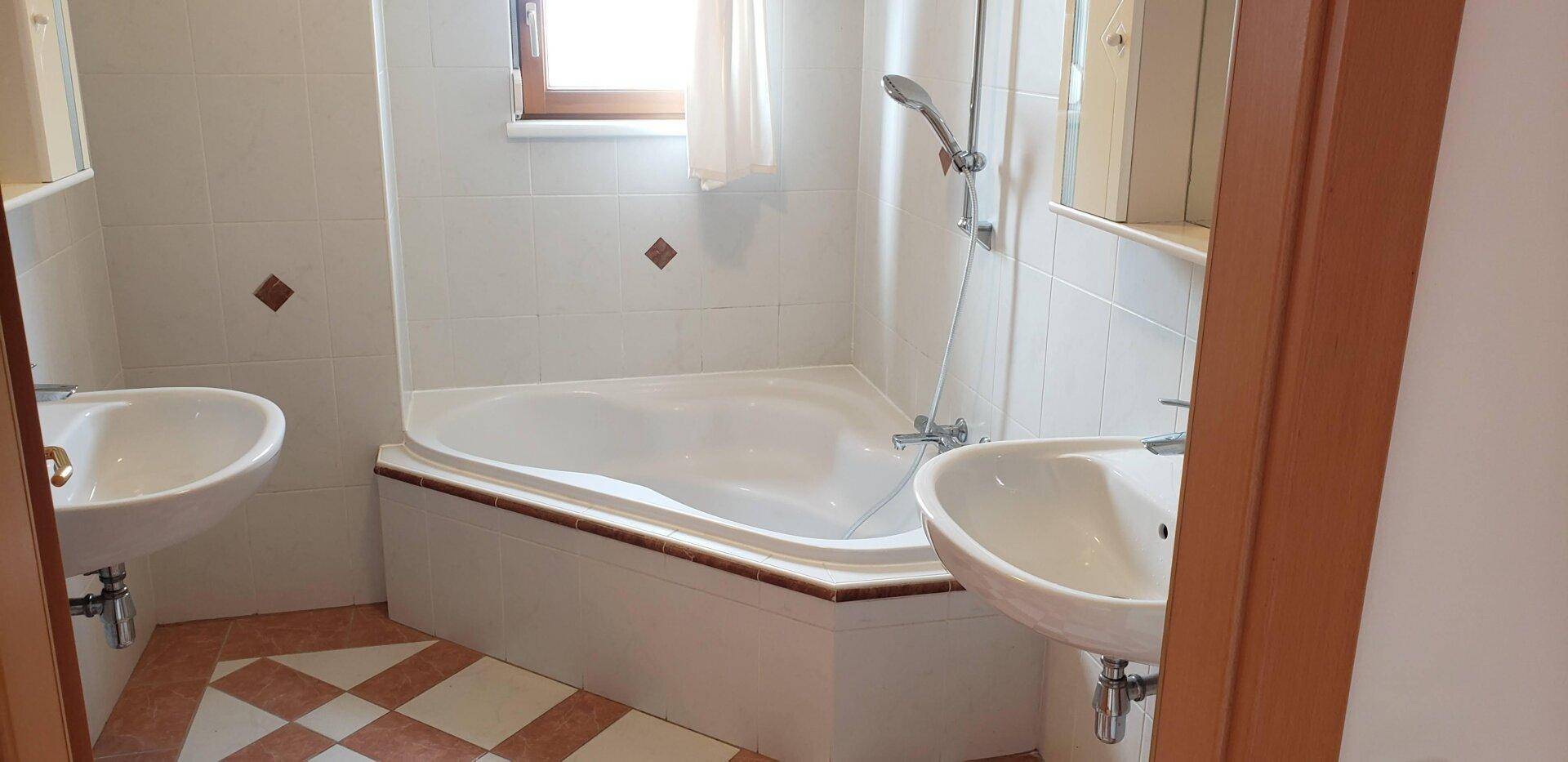 Badezimmer mit Eckbadewanne, zwei Waschbecken und hellen Fliesen, ausgestattet mit Fenster.