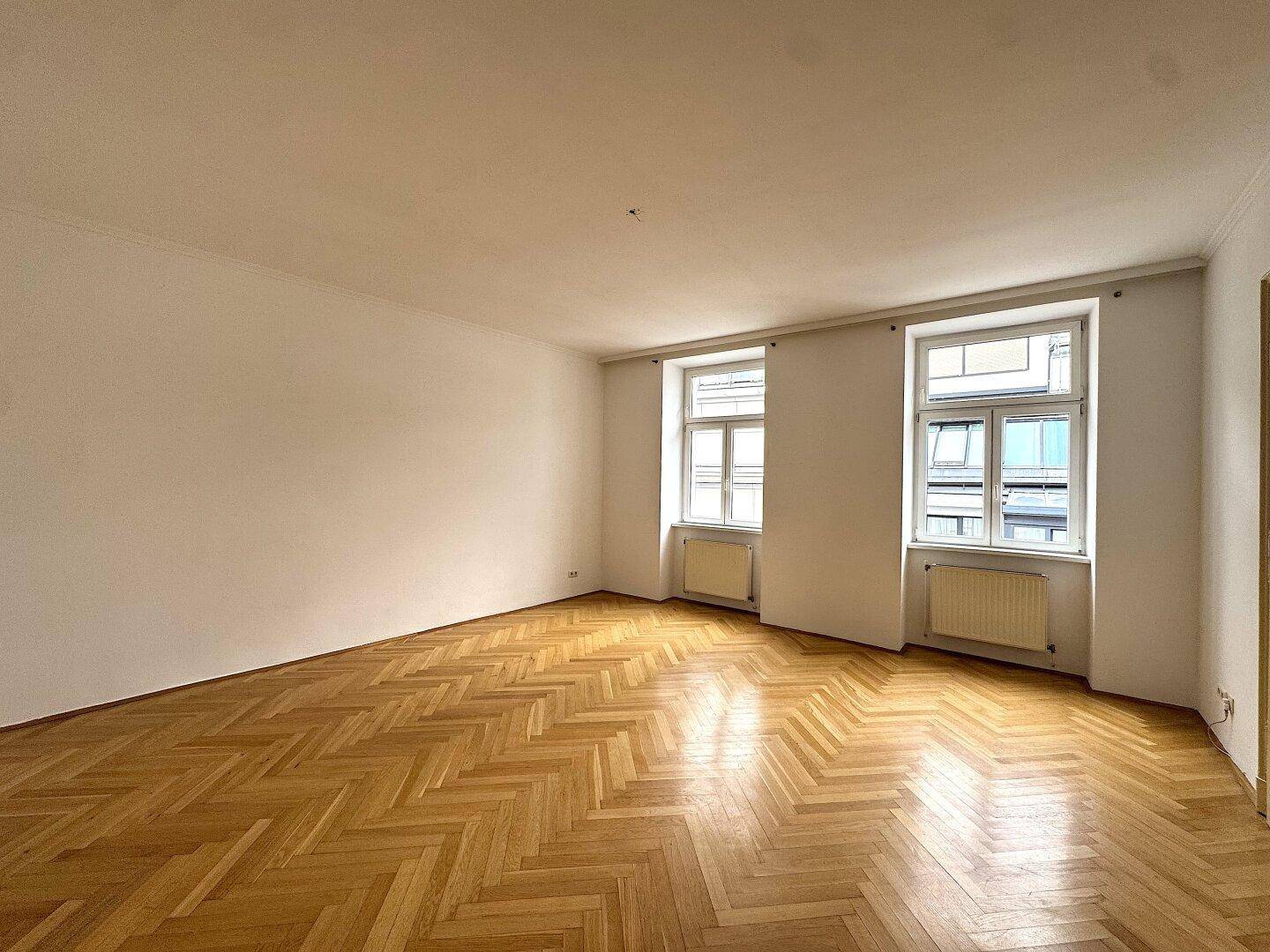 Helles, leeres Zimmer mit klassischem Fischgrätparkett und zwei großen Fenstern.