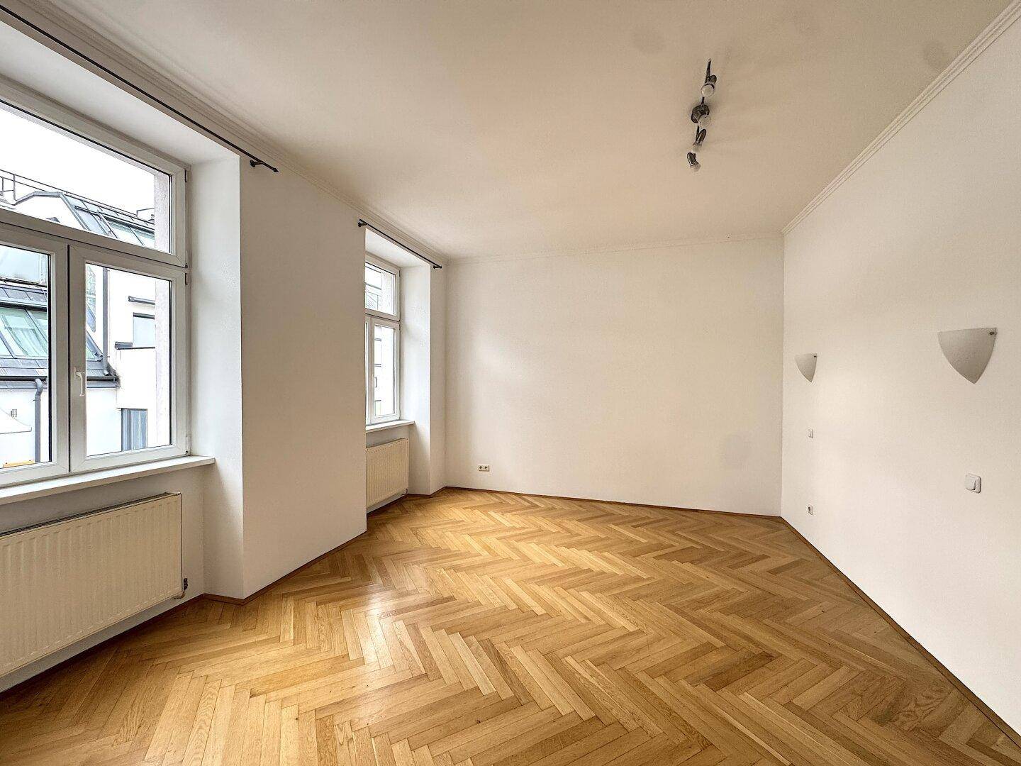 Geräumiges Zimmer mit Fischgrätparkett, hohen Decken und dekorativen Wandleuchten.