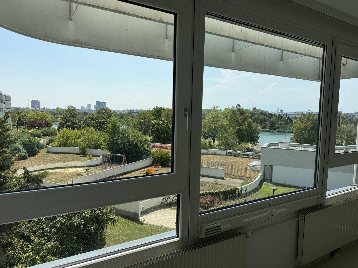 Weitblick aus dem Fenster über einen Garten mit Spielplatz, See und Stadtpanorama.