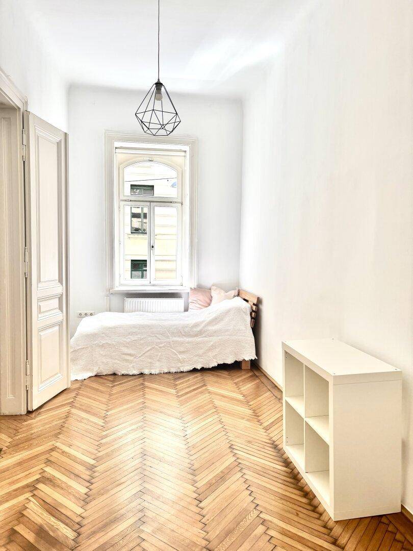 Kleines Schlafzimmer mit Fischgrätparkett, hohen Decken und einem großen Fenster für viel Licht.