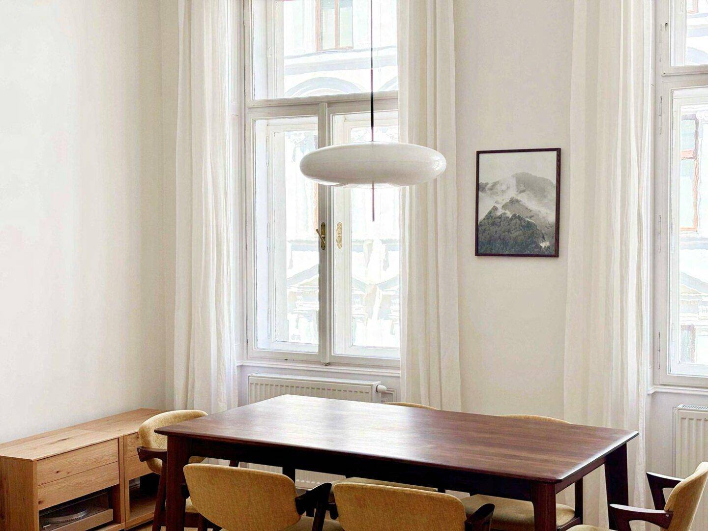 Lichtdurchfluteter Essbereich mit Holztisch, modernen Stühlen und hohen Fenstern mit Vorhängen.